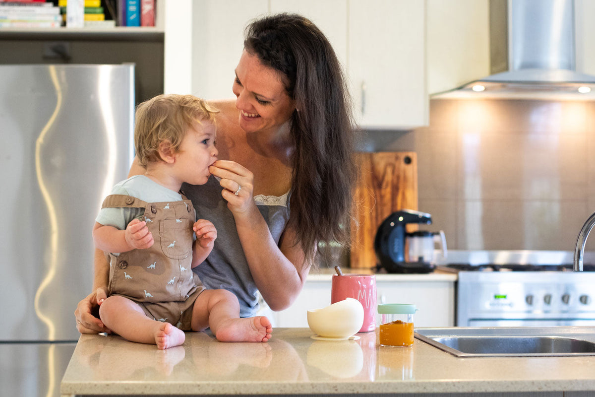 Mother feeding a toddler sitting on a kitchen bench, sharing food in a bright modern kitchen with baby food jar and KiddoKook Pro appliance in the background