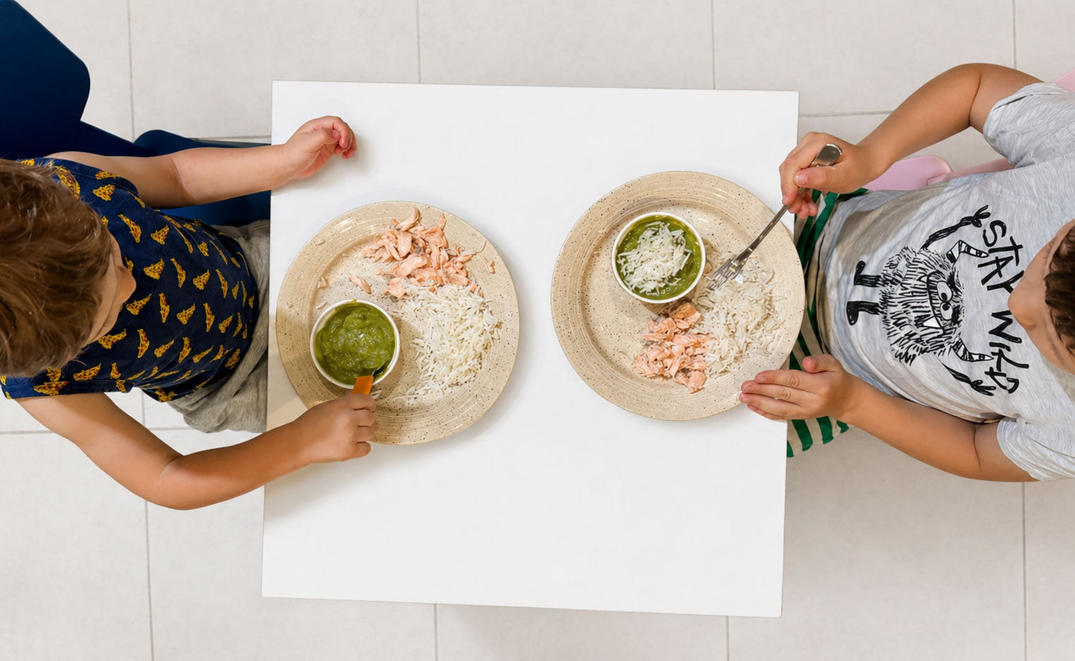 Overhead view of two young children sitting at a small table eating rice, salmon, and green puree, in a bright, clean home setting with soft natural lighting.