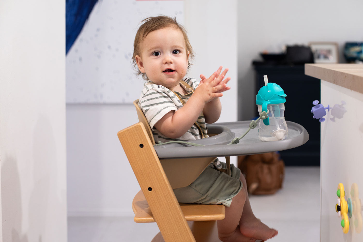 Baby sitting in a high chair clapping hands, with a sippy cup on the tray during mealtime in a home kitchen.