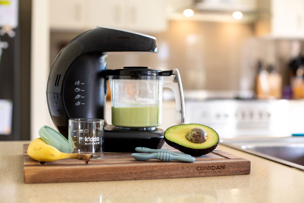 KiddoKook Pro baby food maker blending green puree on a kitchen bench, with avocado, banana, and Kiddo Kitchen jar and spoons in the foreground.