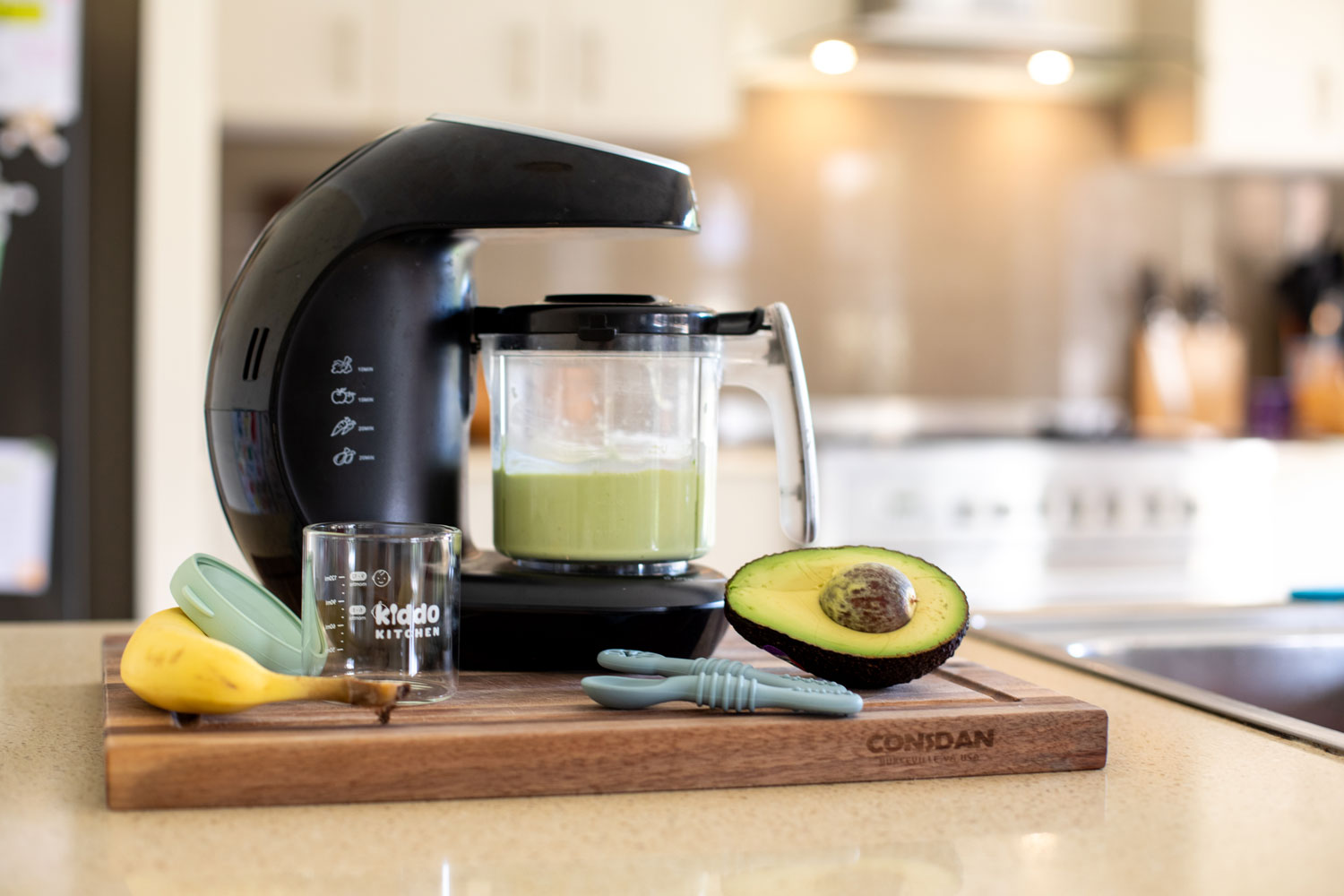 KiddoKook Pro baby food maker blending green puree on a kitchen bench, with avocado, banana, and Kiddo Kitchen jar and spoons in the foreground.